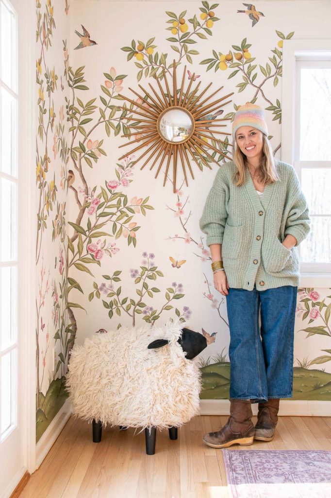 A person in a green cardigan and blue jeans stands in a brightly lit room with floral wallpaper. They wear a hat and boots and smile beside a fluffy sheep-shaped stool. A sunburst mirror is on the wall.