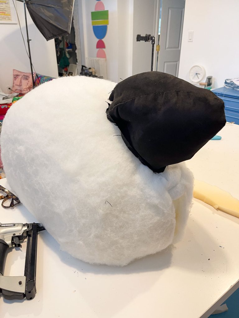 A large white fluffy object with a smaller black section on top rests on a worktable. A stapler and foam pieces are nearby. The background features colorful art supplies and a partially visible canvas.