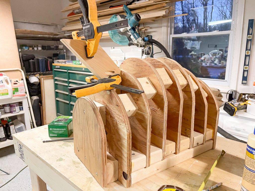 A wooden structure is being built on a workbench using plywood pieces clamped together. Various tools and materials are scattered around, including a saw and a green toolbox. A window shows trees and a vehicle outside.