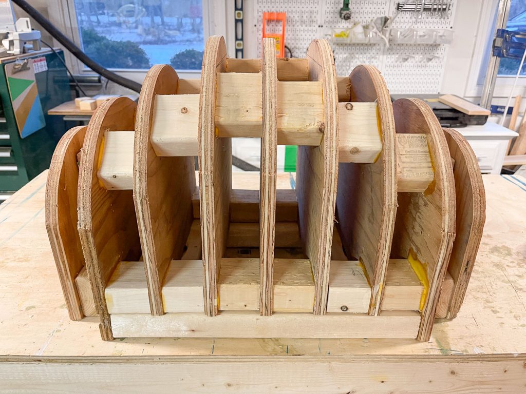 Wooden structure with multiple vertical and horizontal curved panels, resembling a series of arches. The background shows a workshop with various tools and equipment.