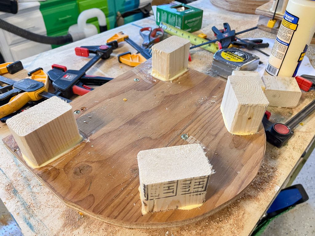 A wooden board with four square wooden blocks attached using glue is placed on a workbench. Various woodworking tools, such as clamps, a tape measure, and glue, are scattered around it.