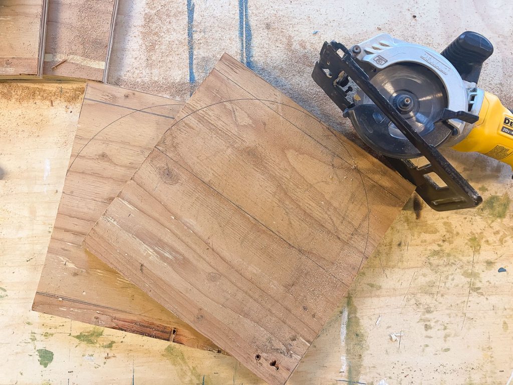 A circular saw rests on a wooden surface next to two square pieces of wood with penciled semi-circle markings. The scene suggests preparations for cutting curved shapes from the wood.