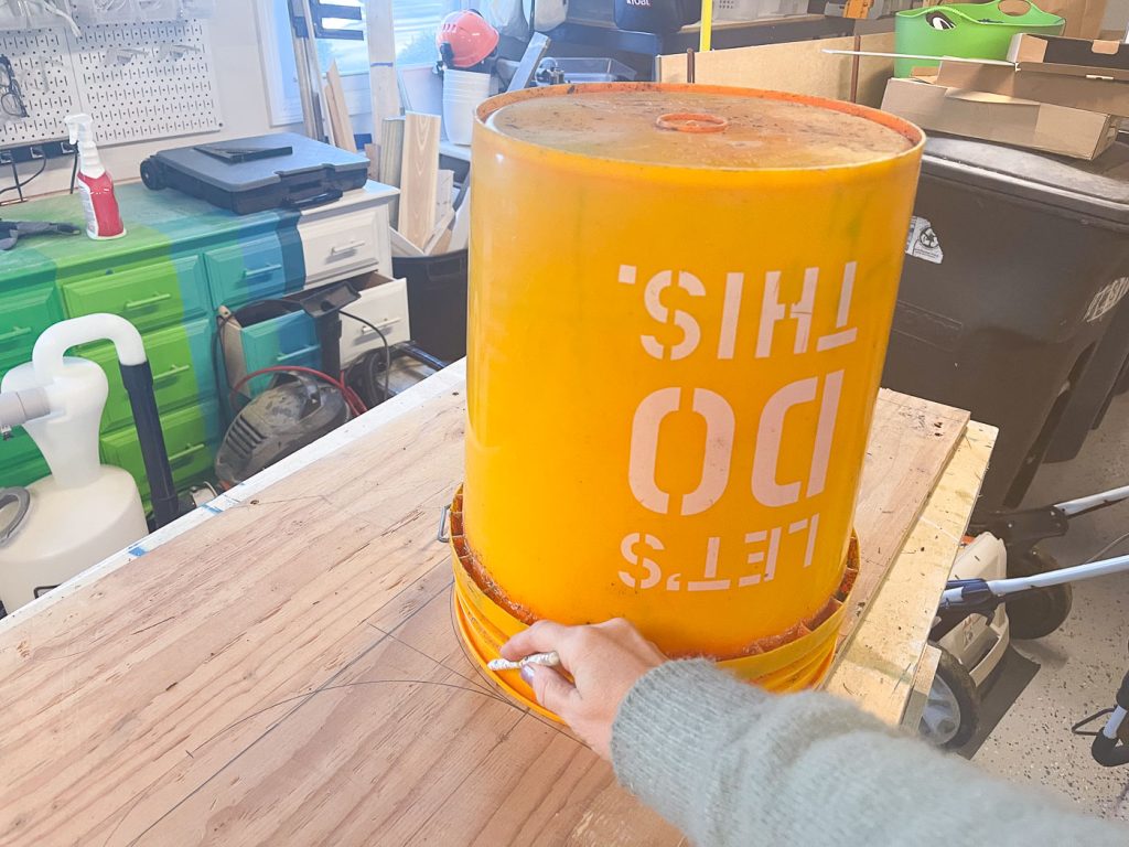 A person is using a yellow bucket as a stencil to draw a semicircle on a wooden surface. The bucket has "LET'S DO THIS" printed on it in white letters. The background shows a workshop with various tools and equipment.