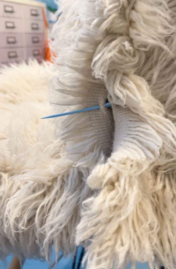 Close-up of a furry, off-white textile with a single blue knitting needle inserted in the middle. In the background, a blurred office setting with drawer units. The texture of the textile is shaggy and layered.