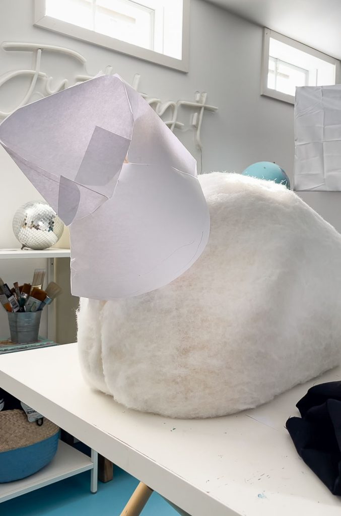 A work in progress of a soft, rounded white object topped with a conical paper structure, possibly resembling a sheep or animal. It rests on a table in a bright room, surrounded by various crafting supplies.