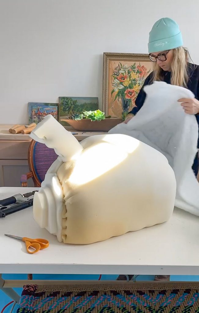 A person wearing a turquoise beanie is standing at a table, wrapping a large foam object with white fabric. Scissors and a staple gun are on the table, and artwork is displayed in the background.