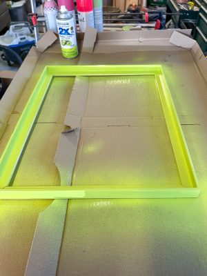 A bright yellow-green picture frame being spray-painted on a cardboard surface, with several cans of spray paint in the background.