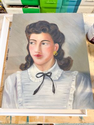A portrait painting of a woman with dark hair styled in waves. She wears a white ruffled blouse with a black ribbon tied at the collar and gold hoop earrings. The painting rests on a wooden surface with colorful drawers visible in the background.