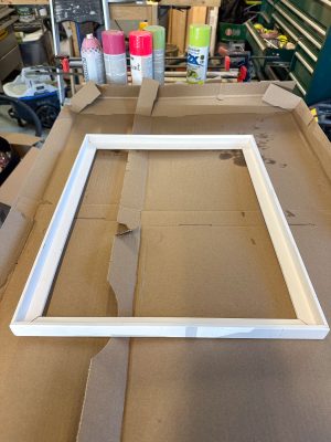 A white frame on a cardboard surface in a workshop setting, surrounded by various spray cans and tools in the background.
