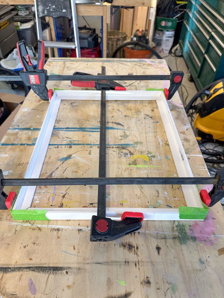 A wooden work table in a cluttered workshop holds a white rectangular frame secured by red and black clamps. Green tape is placed on the corners of the frame. Various tools and equipment are visible in the background.