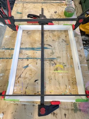 A rectangular wooden frame is clamped securely on a workbench. The surface of the bench is covered in paint splatters and markings. Clamps with red handles hold the frame in place, and a roll of green tape is visible in the background.