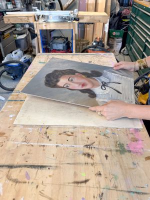 A person, with manicured nails, holds a painted portrait of a woman with brown hair and a white blouse, over a wooden table. The background includes tools and equipment in a workshop setting.