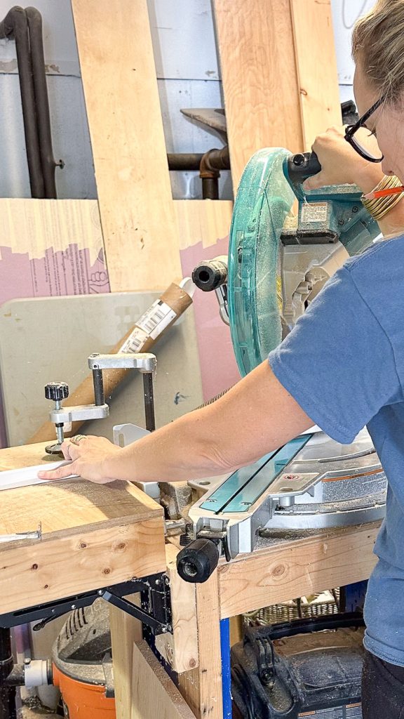 A person using a circular saw to cut a piece of wood in a workshop. Safety goggles are on their head, and they are surrounded by various tools and wooden boards.