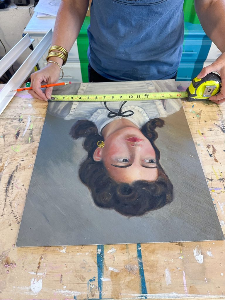 A person measures a portrait painting on a wooden table using a tape measure. The painting, featuring a woman with dark hair and yellow earrings, is positioned upside down. The person wears a blue shirt and gold bracelets.