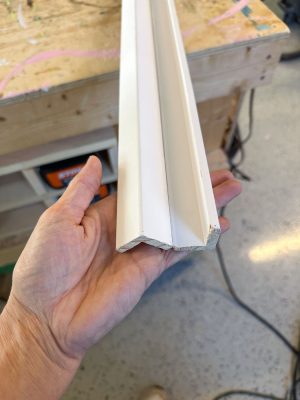 A hand holds a piece of white crown molding with an angled design. The molding is displayed against a wooden workbench in a workshop setting. The floor is visible with some tools and materials around.