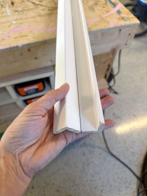 A hand holds two sections of white crown molding against a workbench. The molding ends are displayed to show their profiles, with tools and workshop equipment faintly visible in the background.