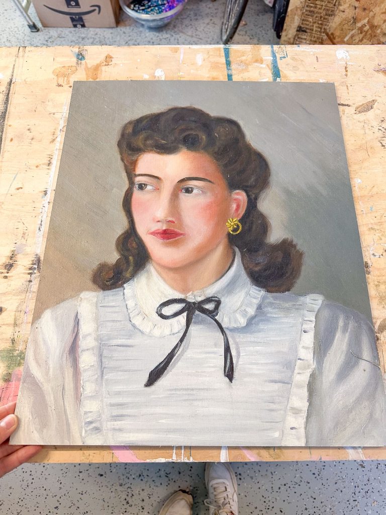 A portrait painting of a woman with wavy dark hair wearing a white ruffled blouse and a black ribbon. She has gold earrings and is gazing to the side. The portrait is set on a wooden surface with a hand holding it.