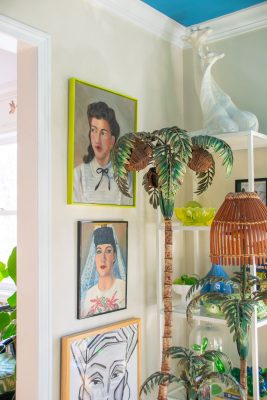 A colorful room with framed portraits on the wall, a sculpture of a peacock on a shelf, and an artificial palm tree beside a lamp. The decor includes plants and various decorative items.