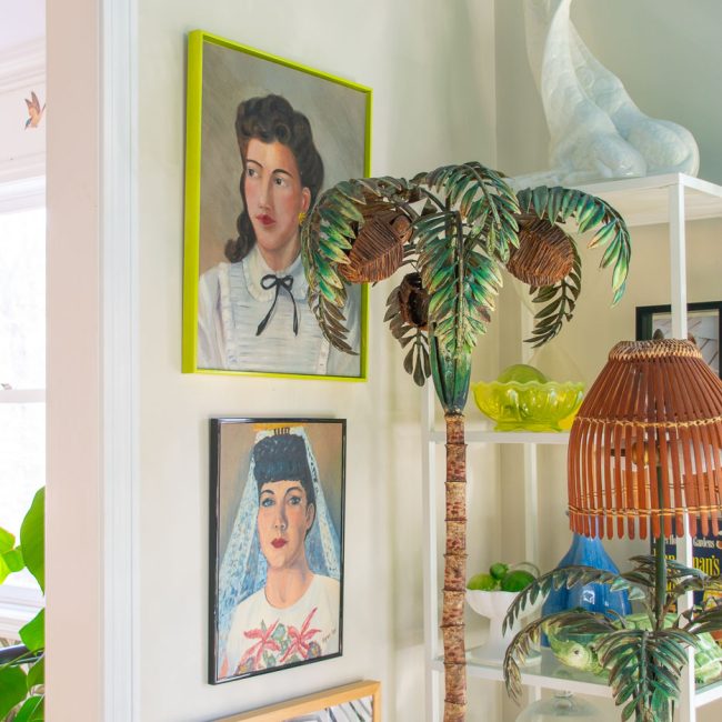 A colorful room with framed portraits on the wall, a sculpture of a peacock on a shelf, and an artificial palm tree beside a lamp. The decor includes plants and various decorative items.