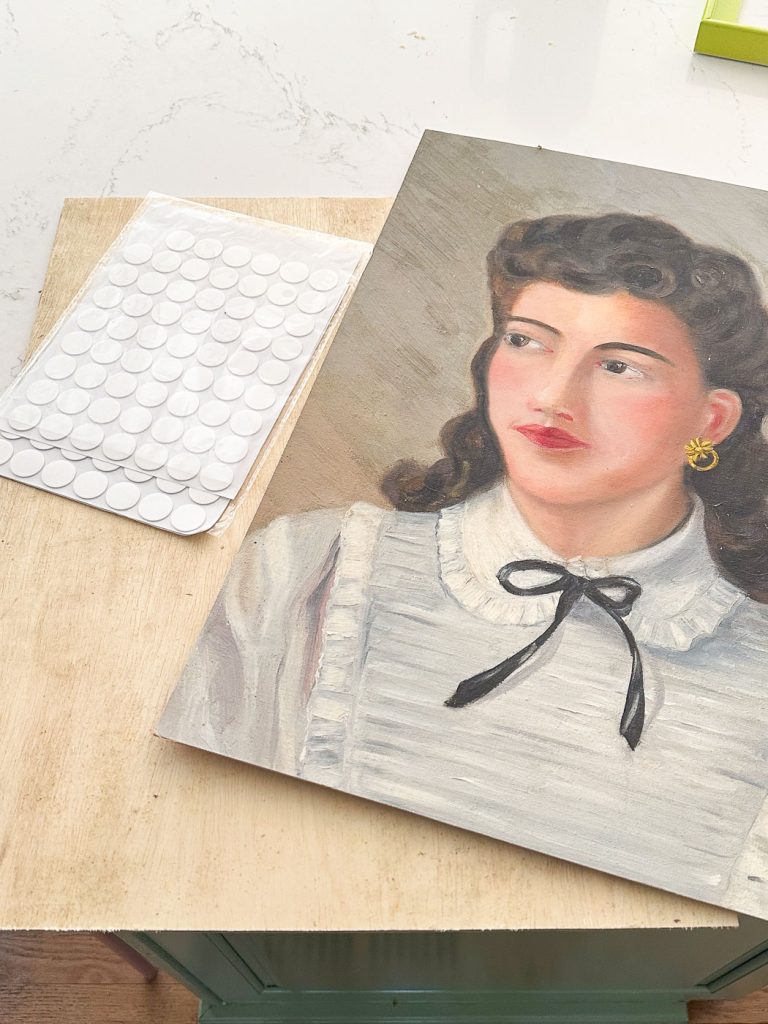 A portrait painting of a woman with wavy dark hair, wearing a white blouse with ruffles and a black ribbon bow, is placed on a wooden surface next to a sheet of adhesive round tabs.