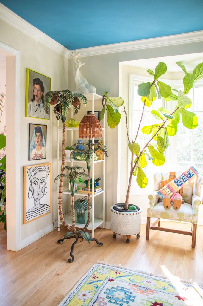 A vibrant room with a blue ceiling and light wooden floor. A large potted plant stands beside a tall lamp with a wicker shade. The walls feature eclectic artwork. A cushioned armchair with colorful pillows sits by a window, next to a small shelf.