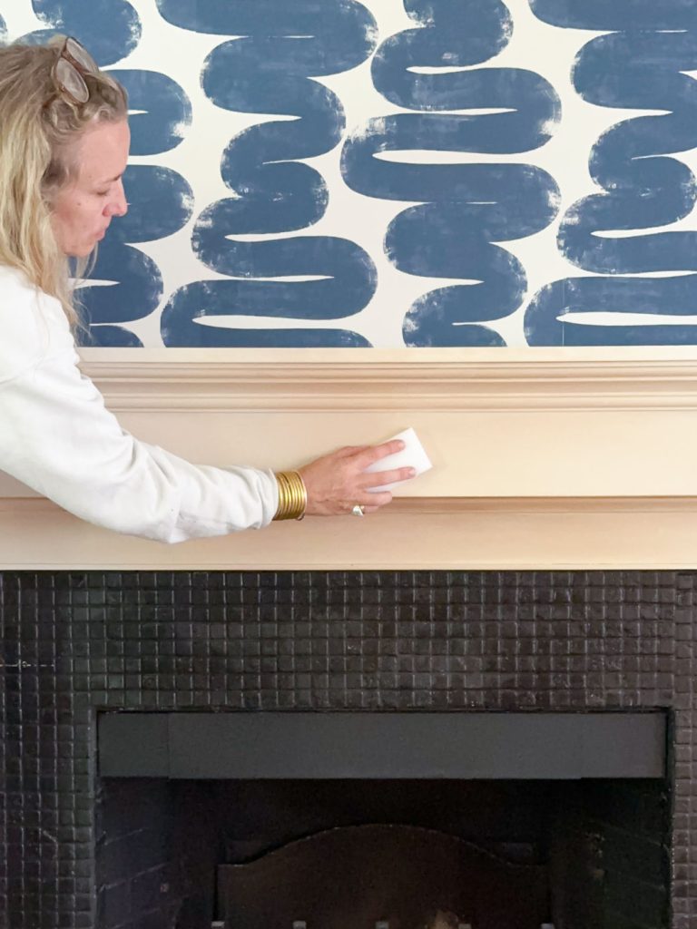 A person wearing a white sweater is cleaning a wooden mantel above a black fireplace with a patterned blue and white wall in the background.