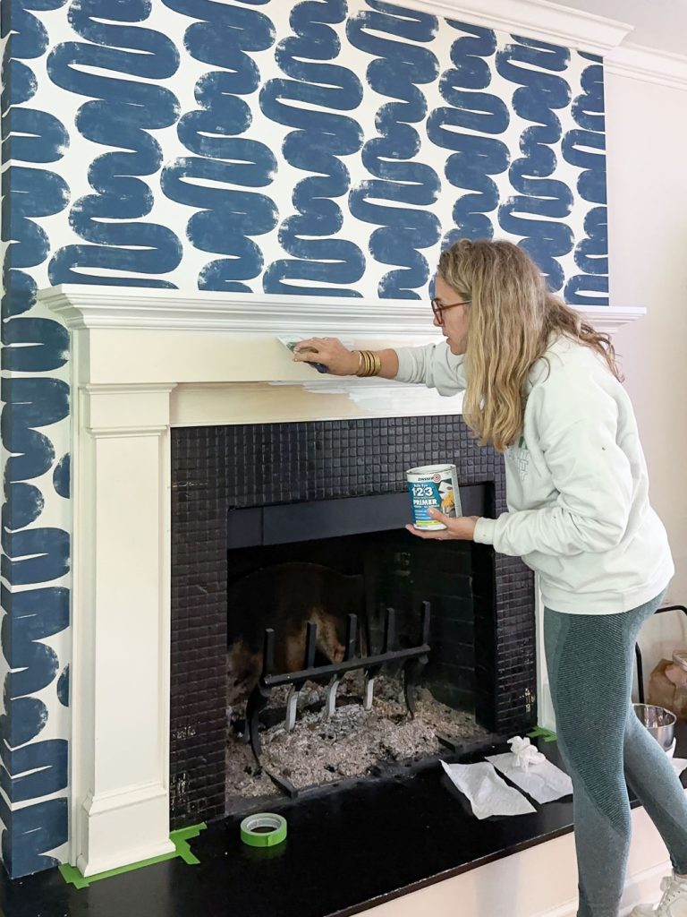 A woman with long hair and glasses paints the white mantel of a fireplace, surrounded by a bold blue abstract design on the wall. She holds a container of primer and a brush, focusing on her work. Tape and cloths are on the floor.