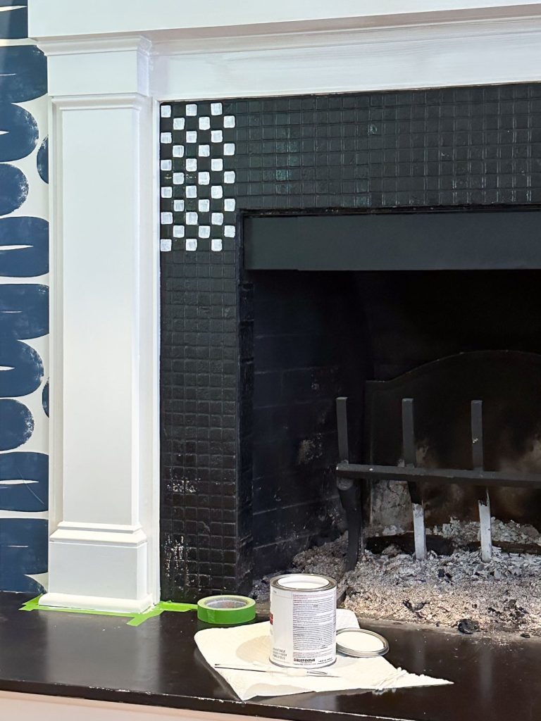 A fireplace being renovated, featuring a partially painted tile design. Black tiles are painted with small white squares in a corner pattern. A can of paint, brush, and cloth are placed nearby on the mantel. Blue wallpaper is visible on the left.