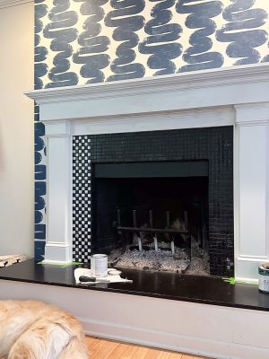 A fireplace with a black tiled interior and white mantel, featuring a unique patterned wallpaper on the wall above. A paint can and brush are on the hearth, suggesting ongoing renovations. A golden retriever is lying on the floor in the foreground.