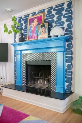 A fireplace with a bright blue mantel and black-and-white checkered tile surround. Above the mantel are two colorful portraits and decorative items. The wall is painted with bold, dark brush strokes. A plant peeks from the left side.