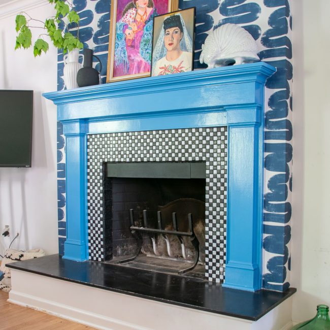 A fireplace with a bright blue mantel and black-and-white checkered tile surround. Above the mantel are two colorful portraits and decorative items. The wall is painted with bold, dark brush strokes. A plant peeks from the left side.