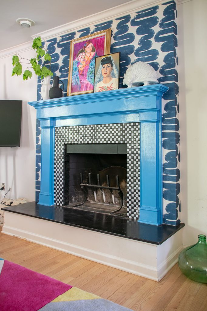 A vibrant living room features a fireplace with a bright blue mantle. Above it, a colorful painting is displayed. The surrounding wall is decorated with bold blue patterns, and a green plant adds a touch of nature to the space. A patterned rug is on the floor.