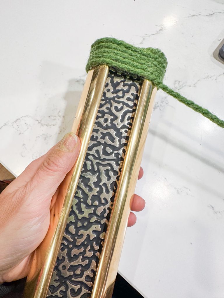 A hand holding an artistic brass and steel object with a Damascus pattern. Green yarn is partially wrapped around the top. The background is a white surface.