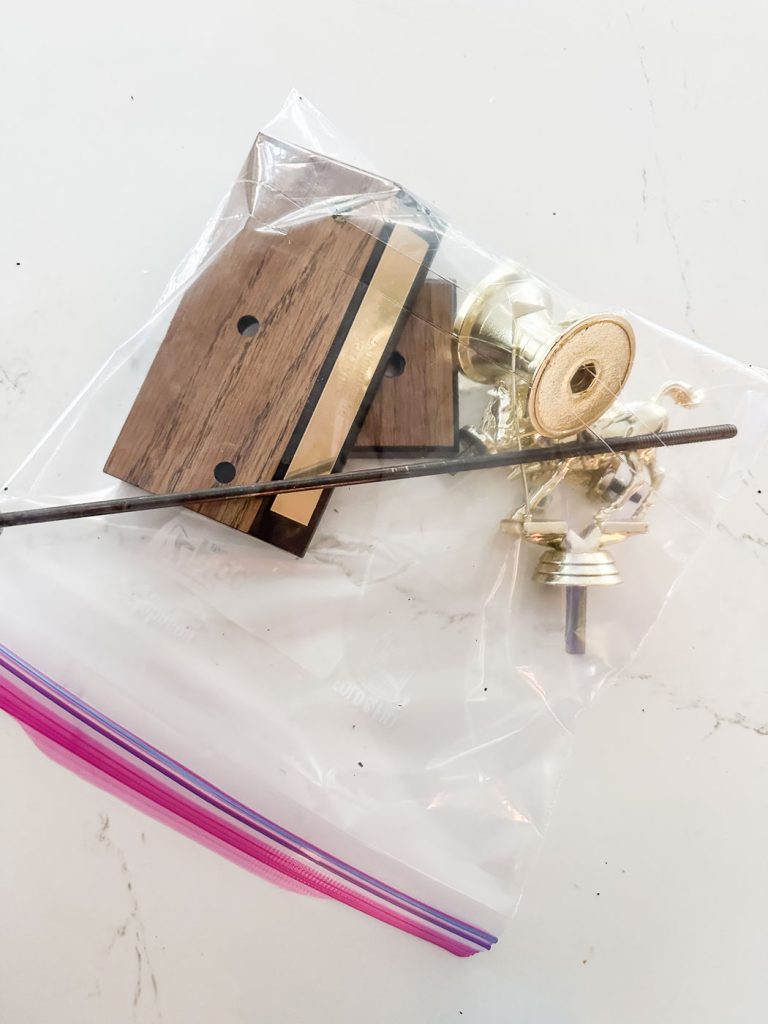 A Ziplock bag containing a brass saxophone neck, a couple of pieces of wood with holes drilled into them, a long metal rod connected to a lock washer and nut and mounted in the bag. They sit on a white surface.