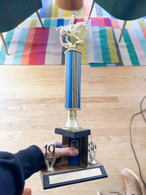 A person is holding a blue and gold trophy with a horse figurine on top. The base features a plaque, and the numbers "1986" are displayed. A colorful striped rug is in the background.