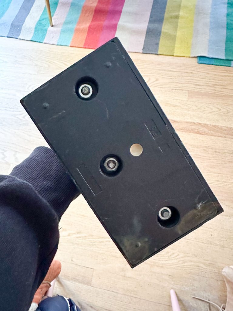 A person holds a black rectangular object with three screw holes and a circular hole in the center. The background shows a wooden floor and a colorful striped rug.