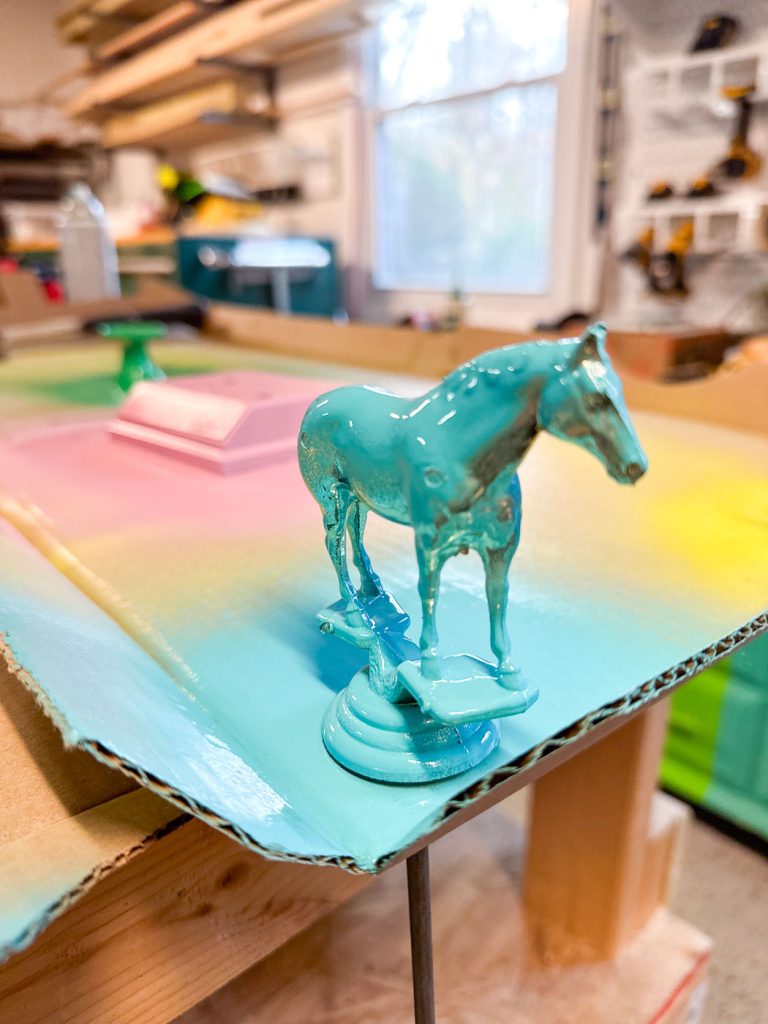 A small horse figurine, painted turquoise, stands on a multicolored spray-painted surface in a workshop. Shelves and tools are visible in the blurred background.