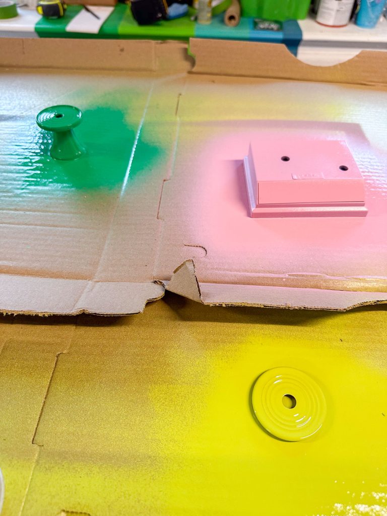 Spray-painted cardboard with a pink wall plate, green drawer knob, and yellow disc spread across. Bright, scattered colors on a textured surface in a workshop setting.