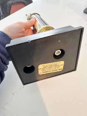A person is holding a gold and silver trophy horizontally by its black base. The base has a yellow label with printed text and two visible screws. The trophy is on a light-colored surface.