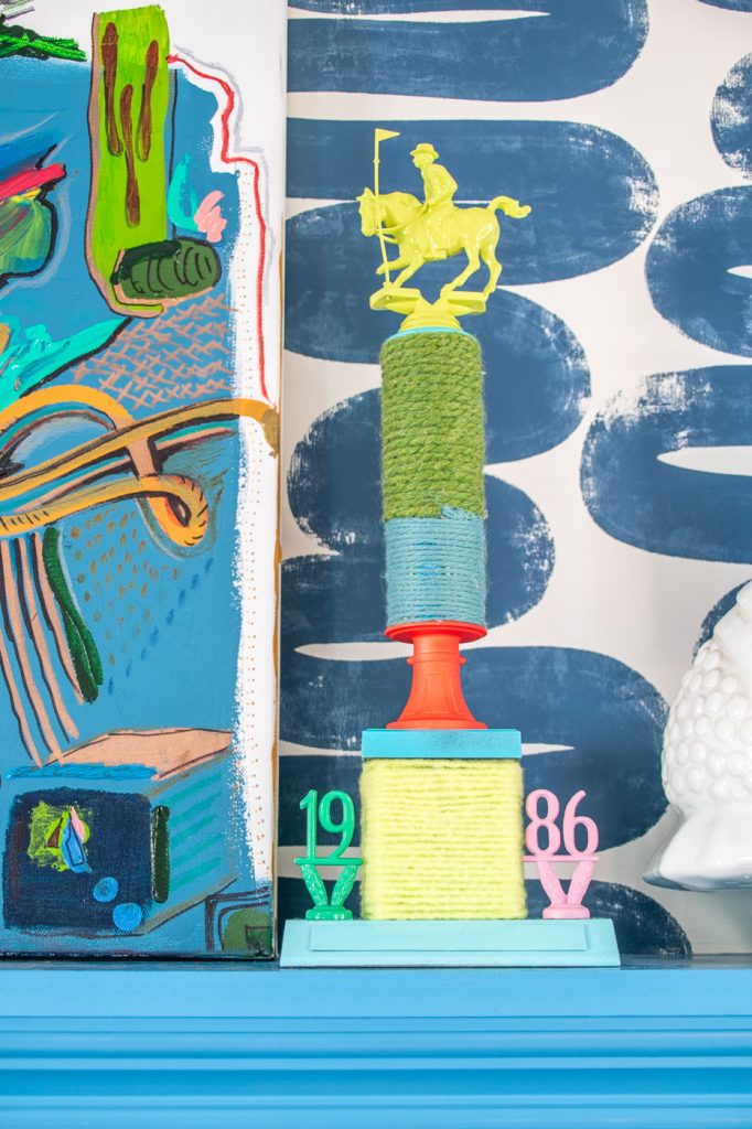 A colorful decorative sculpture featuring a yellow horse and rider atop stacked green and yellow spools. The base is marked with "1986" in pink and numbers in teal. It's set against a vibrant abstract background with blues and greens.