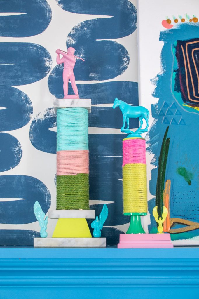 Colorful room decor showing two art pieces on pedestals. Each features wrapped thread spools and miniature figures, including a golfer and a horse. The background has abstract blue and white patterns.