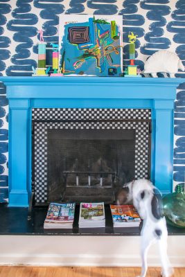 A white and black dog stands in front of a vibrant blue fireplace. The mantel displays colorful art and figurines. The fireplace surround features a black and white checker pattern, and magazines are stacked at its base. Bold, blue wallpaper adorns the wall.