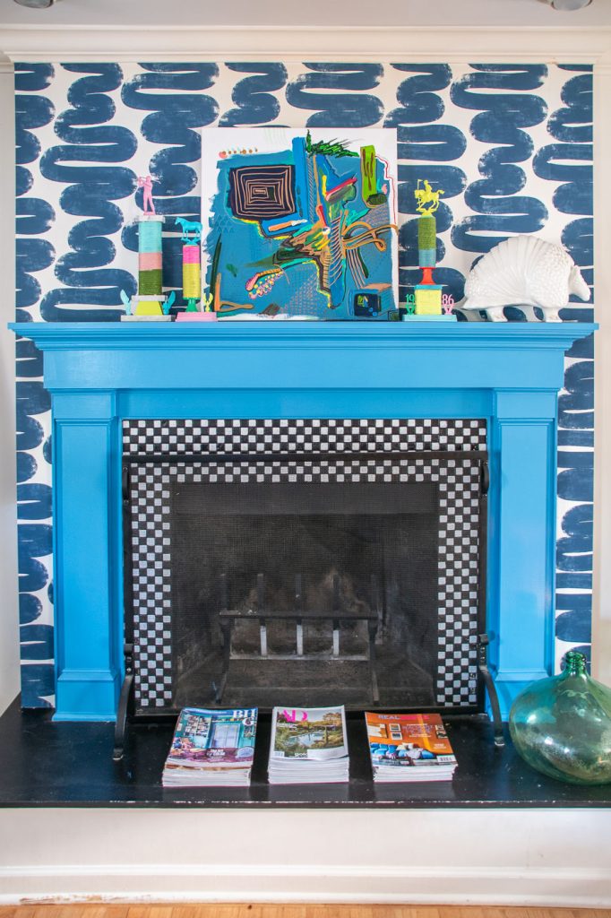 A vibrant blue fireplace with a checkered tile interior is surrounded by abstract wallpaper. The mantel features colorful decorative objects and a piece of abstract art. Magazines are neatly stacked on the floor.