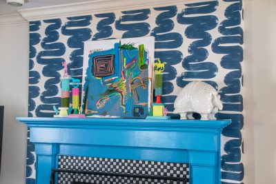 A bright blue fireplace mantel features a colorful abstract artwork and various decorative objects, including a pink figurine and a white shell sculpture, set against a bold blue brushstroke-patterned wall.