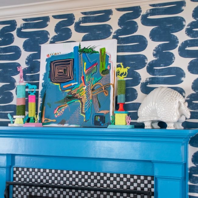 A bright blue fireplace mantel features a colorful abstract artwork and various decorative objects, including a pink figurine and a white shell sculpture, set against a bold blue brushstroke-patterned wall.