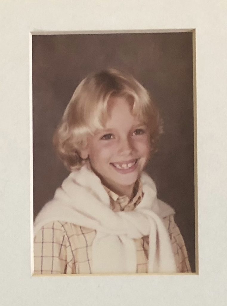 A young child with blonde hair smiles at the camera. They are wearing a plaid shirt and a white sweater draped over their shoulders. The background is a neutral gray tone.