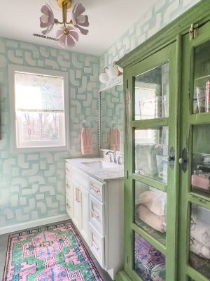 A pastel-themed bathroom with green geometric wallpaper, a green wooden cabinet, and a white vanity. Two windows with floral shades allow natural light. A colorful rug and pink towels add warmth. A unique ceiling light fixture is visible.