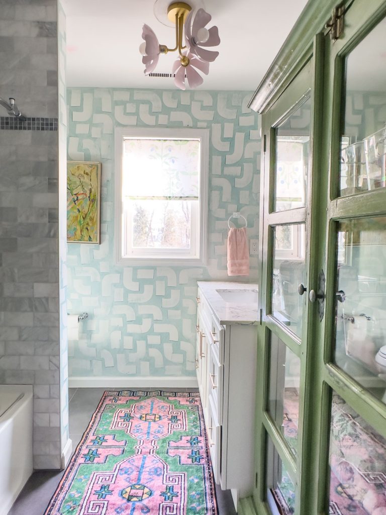 A bathroom with light blue geometric wallpaper, a white vanity, and a green cabinet. A pink and green rug is on the floor, and a window lets in natural light. A floral-shaped ceiling light is above, and a towel hangs on the wall.