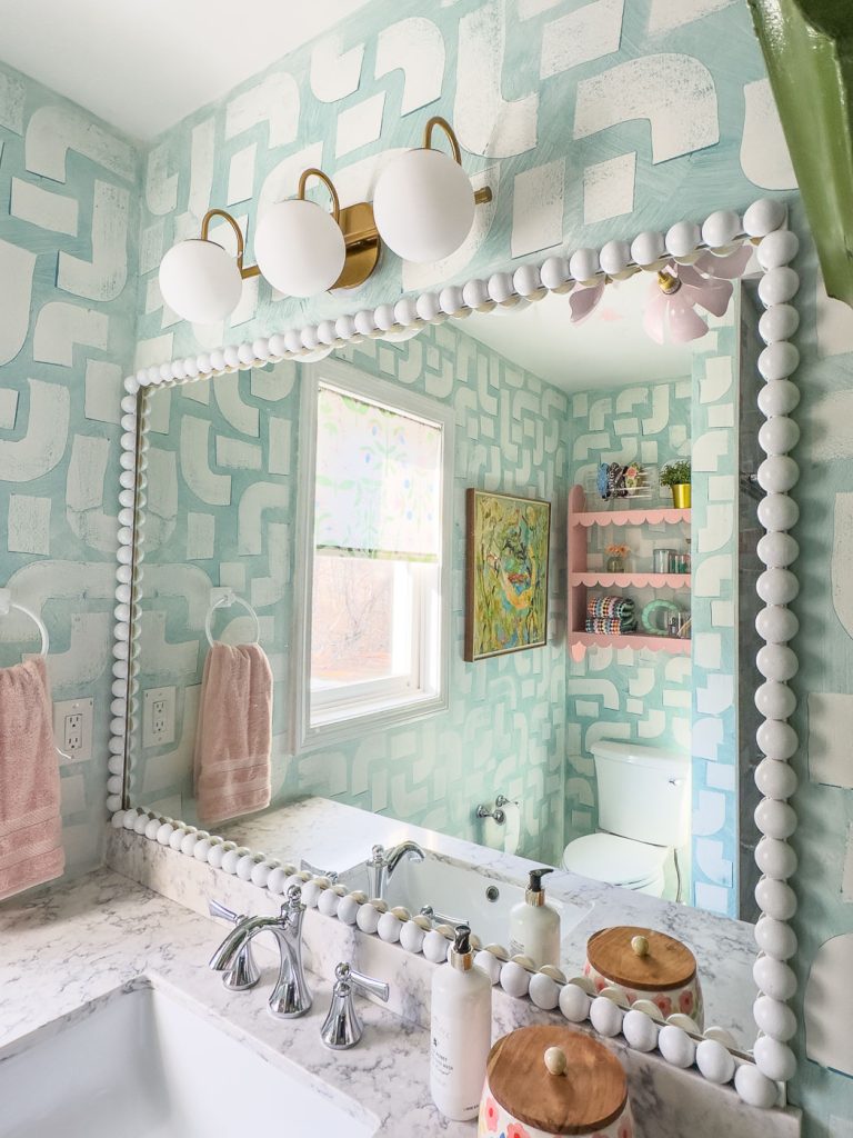 A bathroom with light blue geometric-patterned walls, a large mirror with a white beaded frame, and a marble countertop. There are pink towels, a wall-mounted light fixture with three bulbs, and a shelf with decorative items. A window lets in natural light.