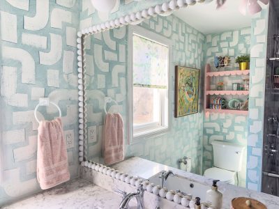 Bathroom with geometric blue and white wallpaper. A large mirror with a beaded frame reflects the room. Marble countertop with a faucet and a lotion bottle. Pink shelves above the toilet hold accessories. A window with a floral blind lets in natural light.